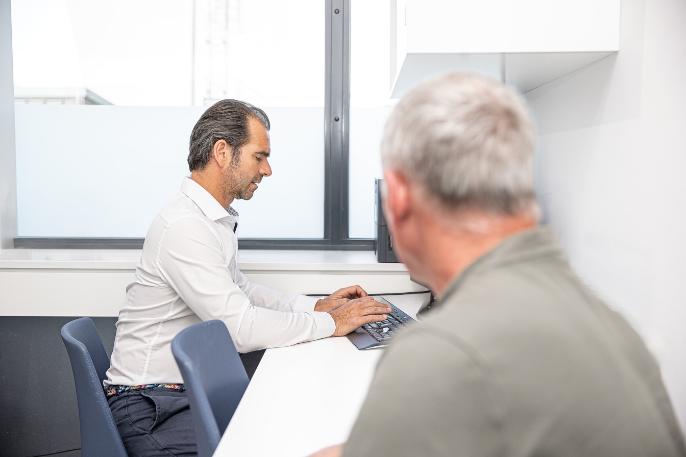 Arzt tippt an einem Computer im Büro, im Vordergrund sitzt eine weitere Person unscharf